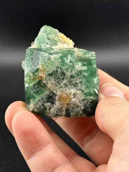 A close-up of a hand holding The Crystalary's LIVE- Winzalite- 12/19/24, a vibrant green mineral specimen against a dark background. This pocket-sized gem features lighter translucent areas with orange and white accents.