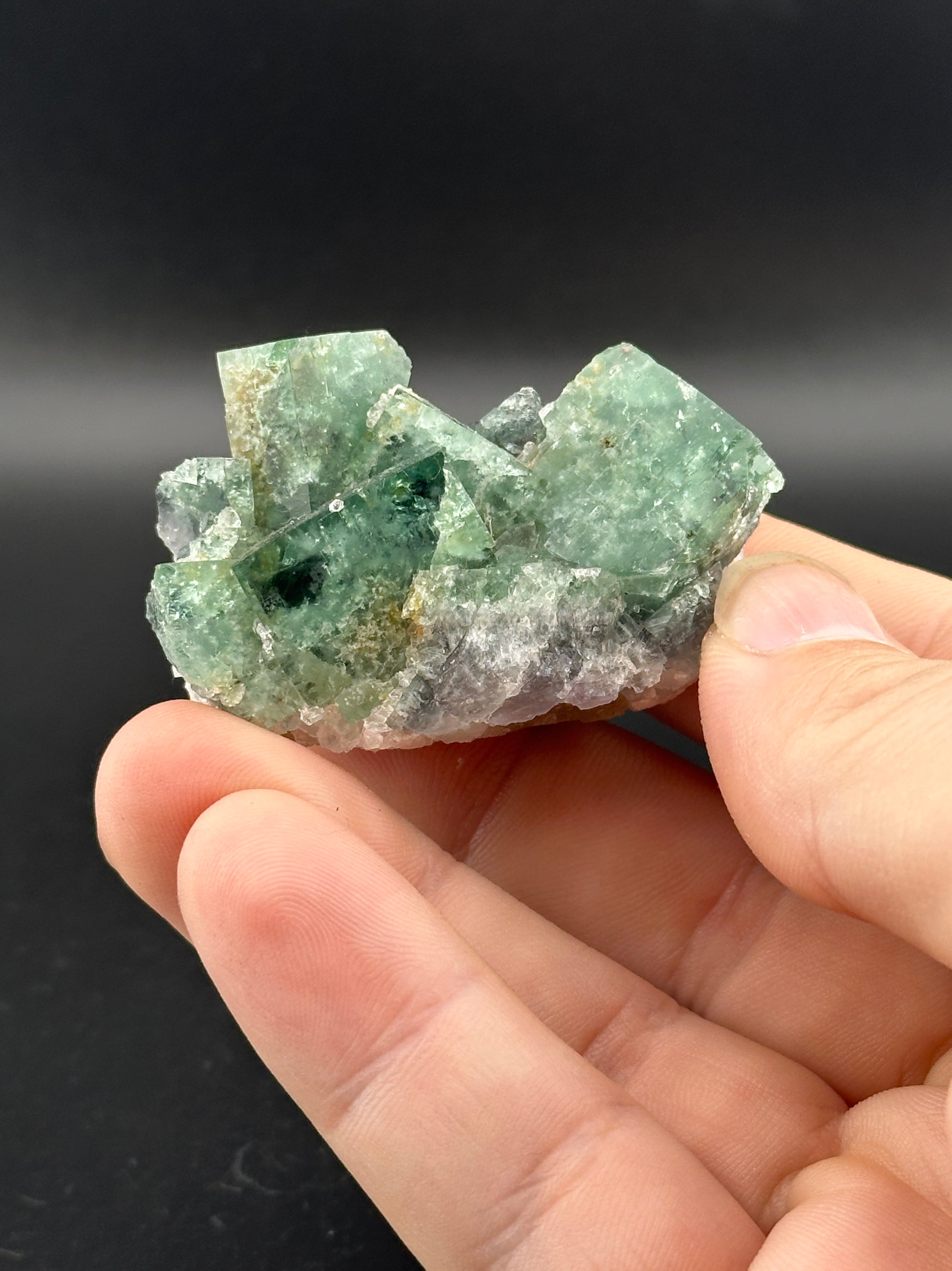 A hand displays The Crystalary's LIVE- Winzalite- 12/19/24, a translucent green fluorite mineral cluster with cubic formations and raw, uneven surfaces against a dark background.