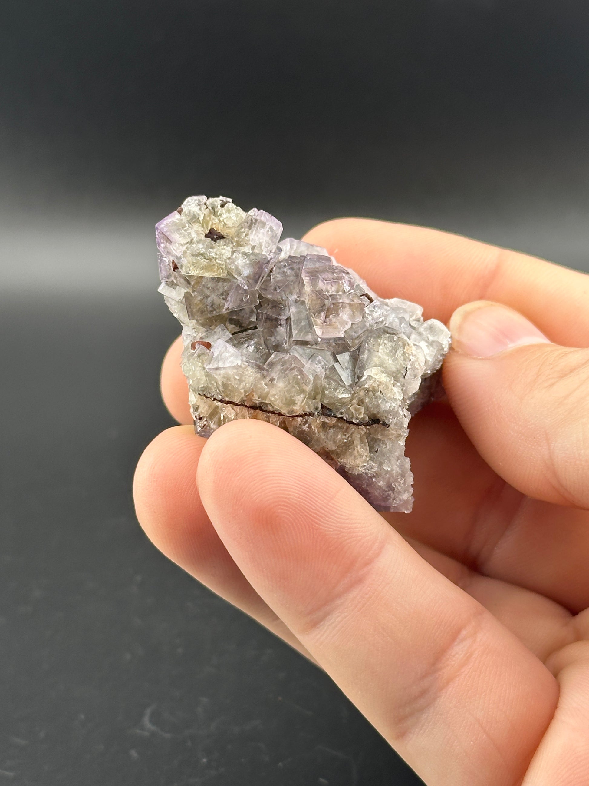 A hand holds a small pocket of Winzalite crystals, part of the LIVE collection from The Crystalary, showcasing clear and purple hues with a slightly rough texture against a dark background.