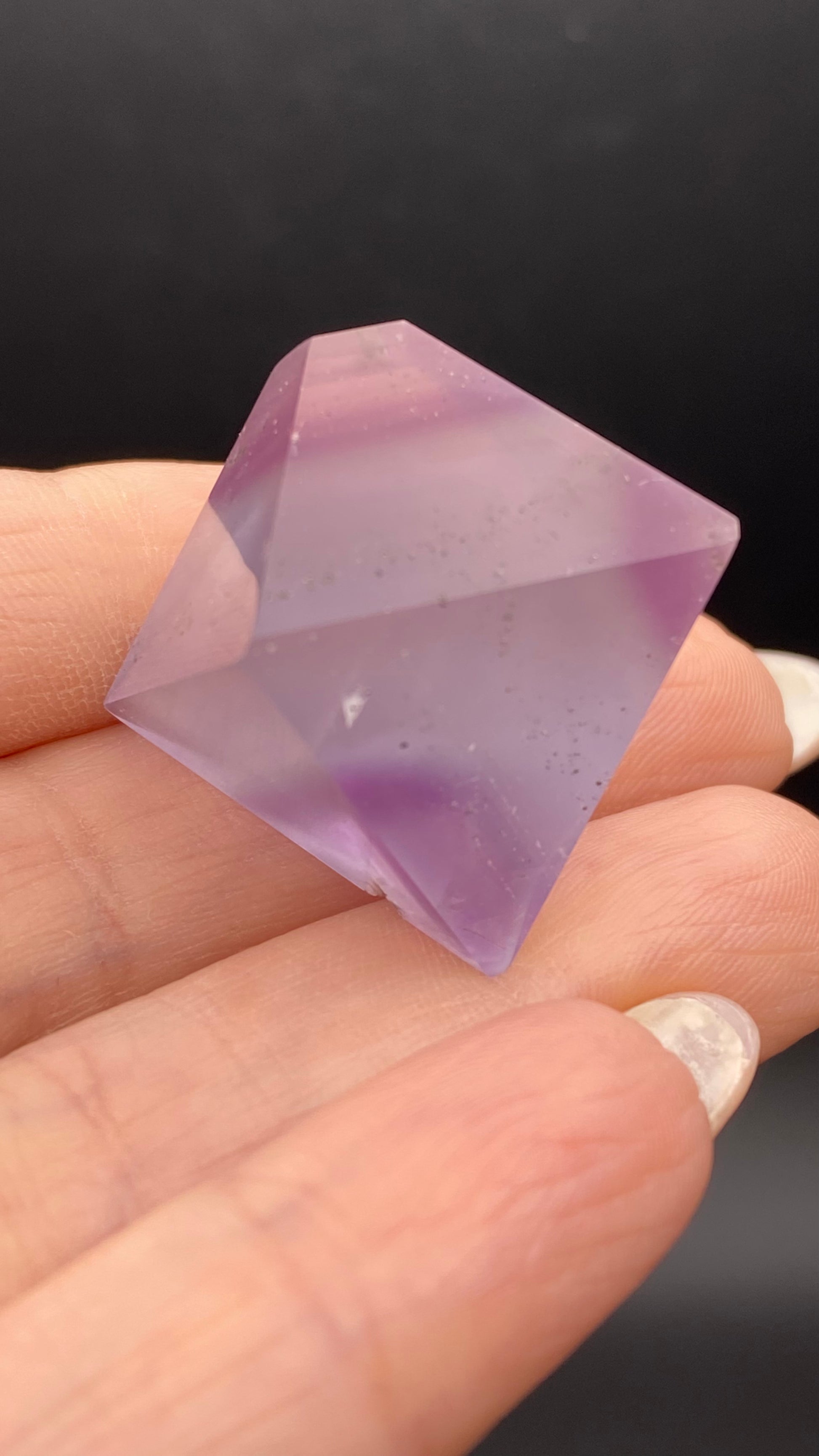 A close-up shot features a manicured hand with glossy nails holding a polished Fluorite Octahedron from Cave-in-Rock, Hardin Co, Illinois against a dark background. This exquisite crystal from The Crystalary boasts facets and transparent layers with subtle internal inclusions visible.