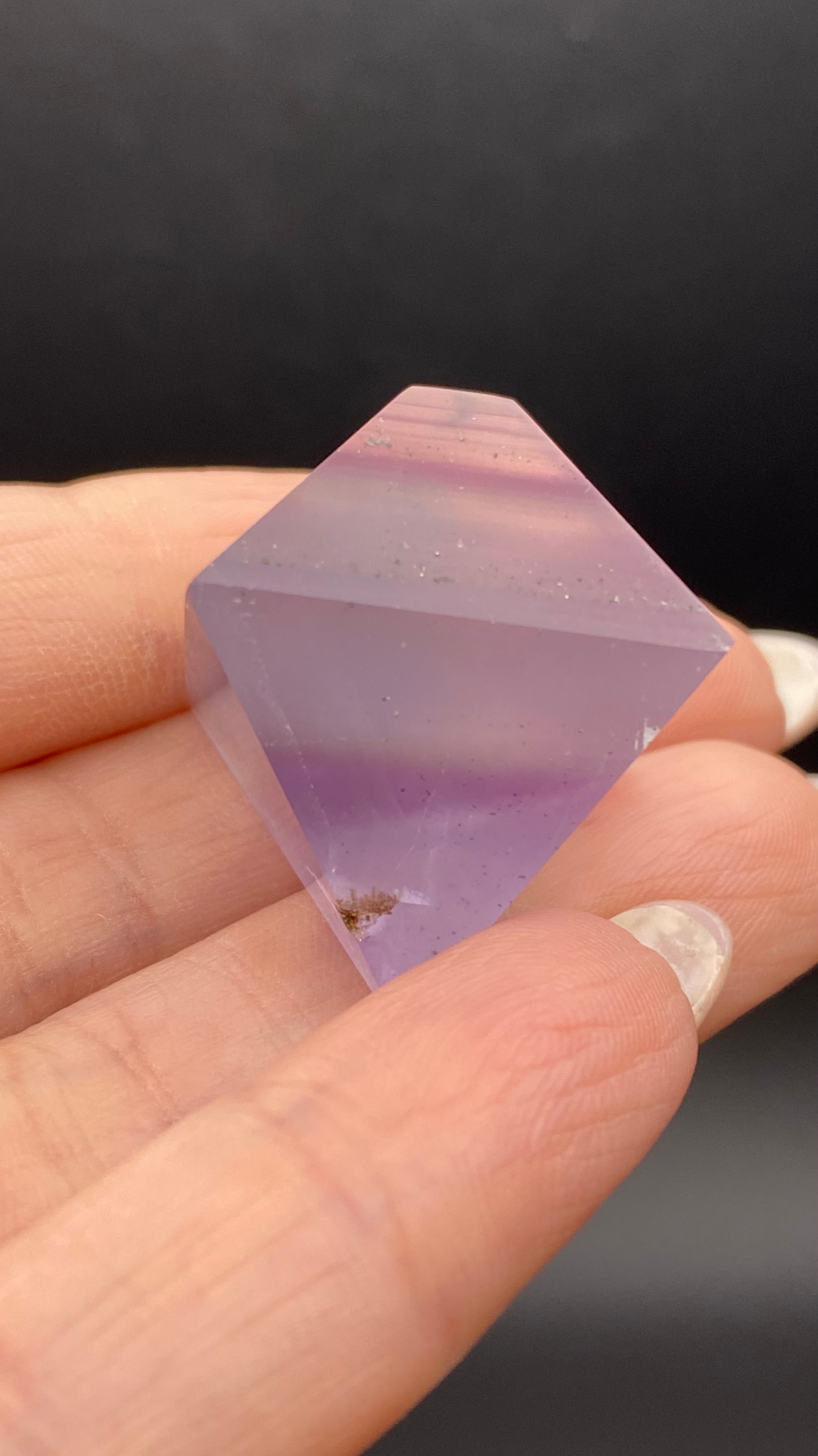 A hand holds The Crystalary's Fluorite Octahedron from Cave-in-Rock, Hardin Co, Illinois. This polished, translucent purple gemstone features an elongated, double-pointed shape with bands of lighter and darker shades along with small inclusions. Set against an out-of-focus background that beautifully emphasizes its intricate patterns.