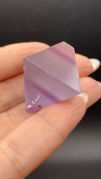 A hand holds a polished, translucent, purple Fluorite Octahedron from The Crystalary, showcasing visible bands of different shades of purple with hints of white. Sourced from Cave-in-Rock in Hardin Co, Illinois, this gemstone catches the light beautifully. The dark background emphasizes the intricate patterns and clarity of the piece.