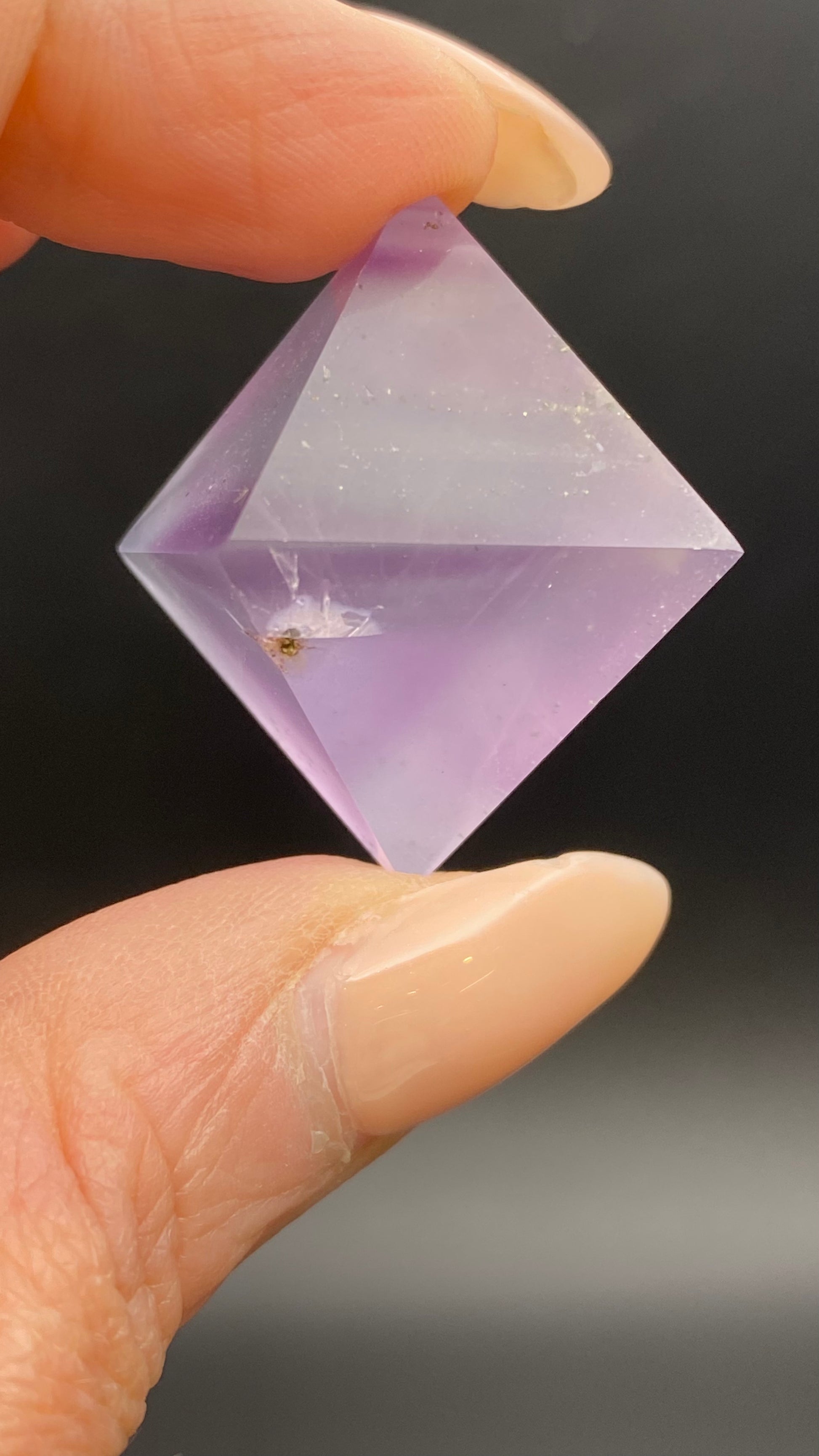 A close-up of a person holding a polished, purple, octahedral crystal from The Crystalary's Fluorite Octahedron - Cave-in-Rock, Hardin Co., Illinois collection between their fingers. The crystal features a small dark inclusion inside it. The background is a blurred dark gray.