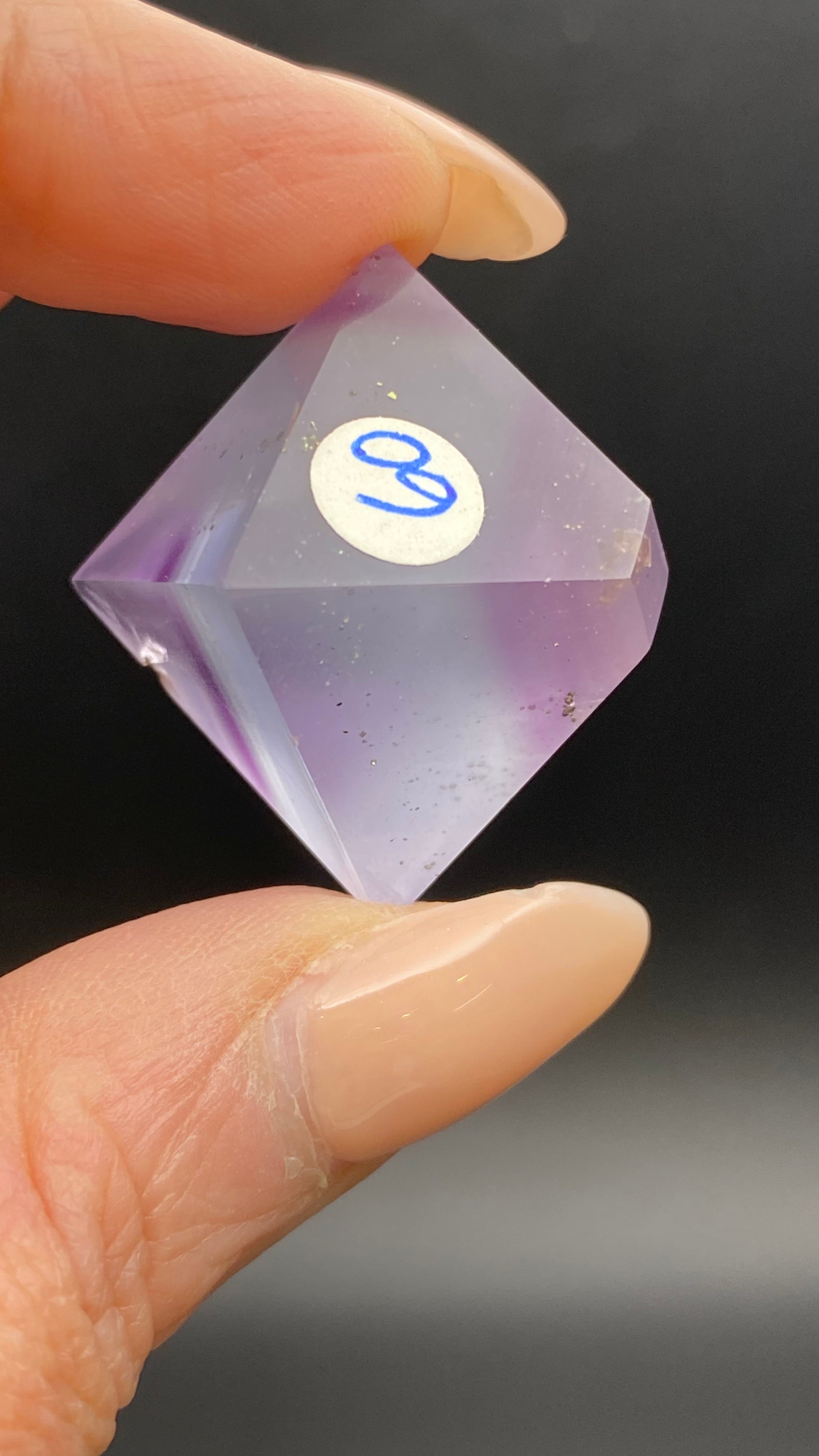 A close-up image of a hand holding a transparent purple Fluorite Octahedron from Cave-in-Rock, Hardin Co, Illinois branded by The Crystalary, with a "30" sticker on it. The fingers have a nude-colored manicure. The background is blurred and dark, highlighting the hand-polished octahedron.