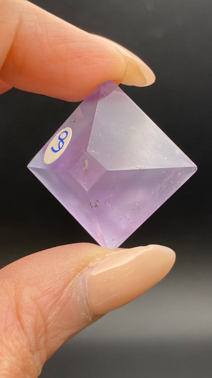 A close-up image of a hand holding a Fluorite Octahedron from Cave-in-Rock, Hardin Co, Illinois by The Crystalary. The crystal is purple and transparent with a double pyramid shape, possibly hand-polished. It features a small, round white sticker with the number "00" on it. The nails are neatly manicured and painted in a neutral color against the dark background.