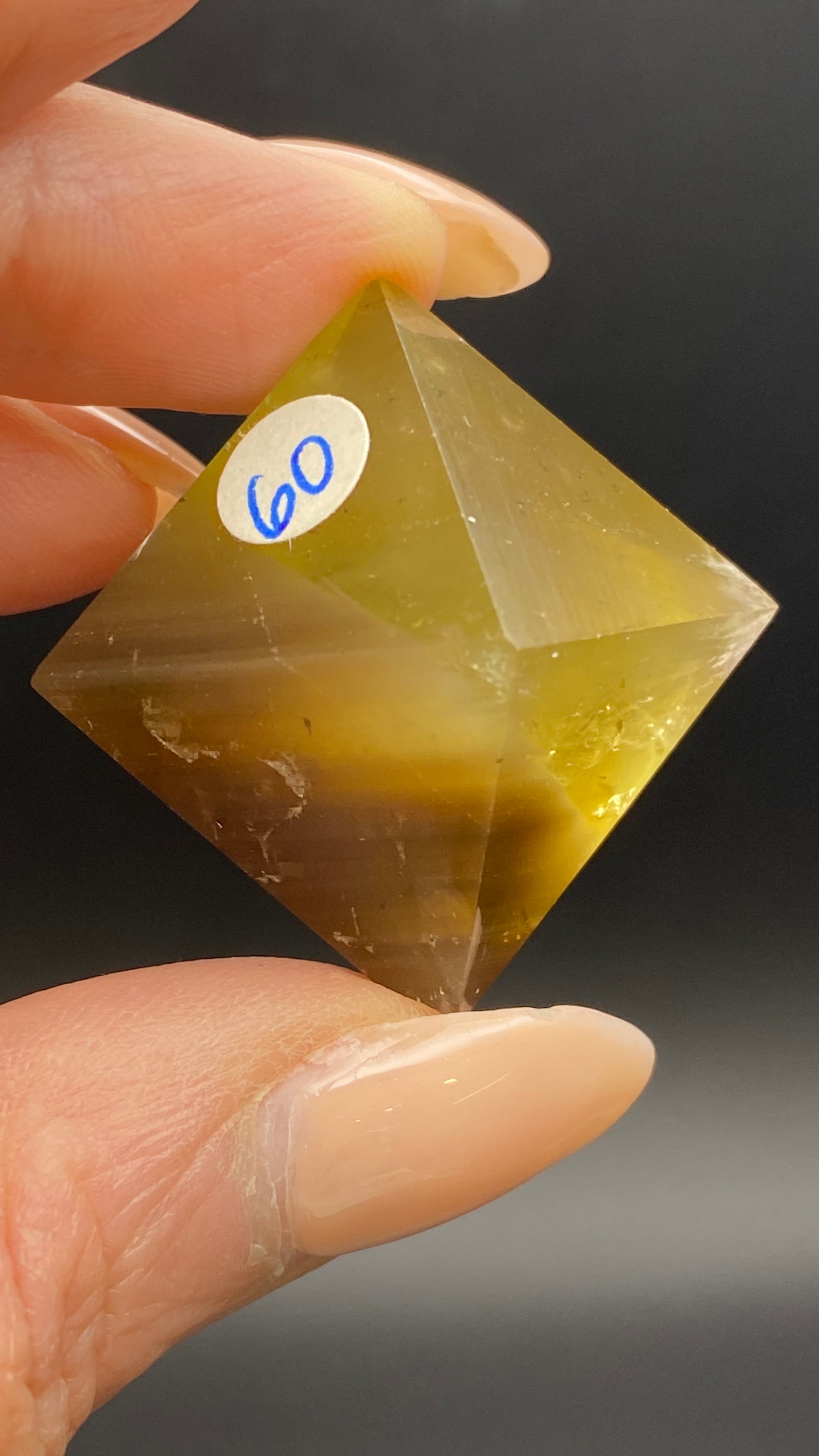 A close-up image of a person's hand holding a yellow-green, hand-polished Fluorite Octahedron- Cave-in-Rock, Hardin Co, Illinois from The Crystalary. The gemstone is meticulously cut into an octahedron shape and features a sticker labeled "60" on one of its faces. The dark background accentuates the vibrant color and intricate facets of the Hardin Co Illinois octahedron.