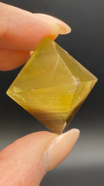 A hand with manicured nails holds a translucent, yellow-brown fluorite octahedron from Cave-in-Rock, Hardin Co, Illinois against a black background. This hand-polished piece from The Crystalary is faceted, showcasing its reflective surfaces and unique internal striations.