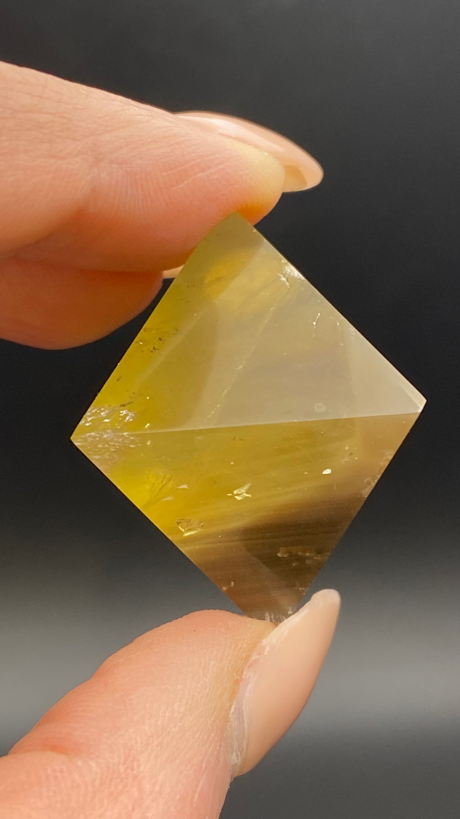 Close-up of a hand holding a geometric, transparent yellow crystal with a combination of angled and flat surfaces. This Fluorite Octahedron from The Crystalary, sourced from the famous Cave-in-Rock in Hardin Co, Illinois, has visible inclusions against a dark, blurred background.