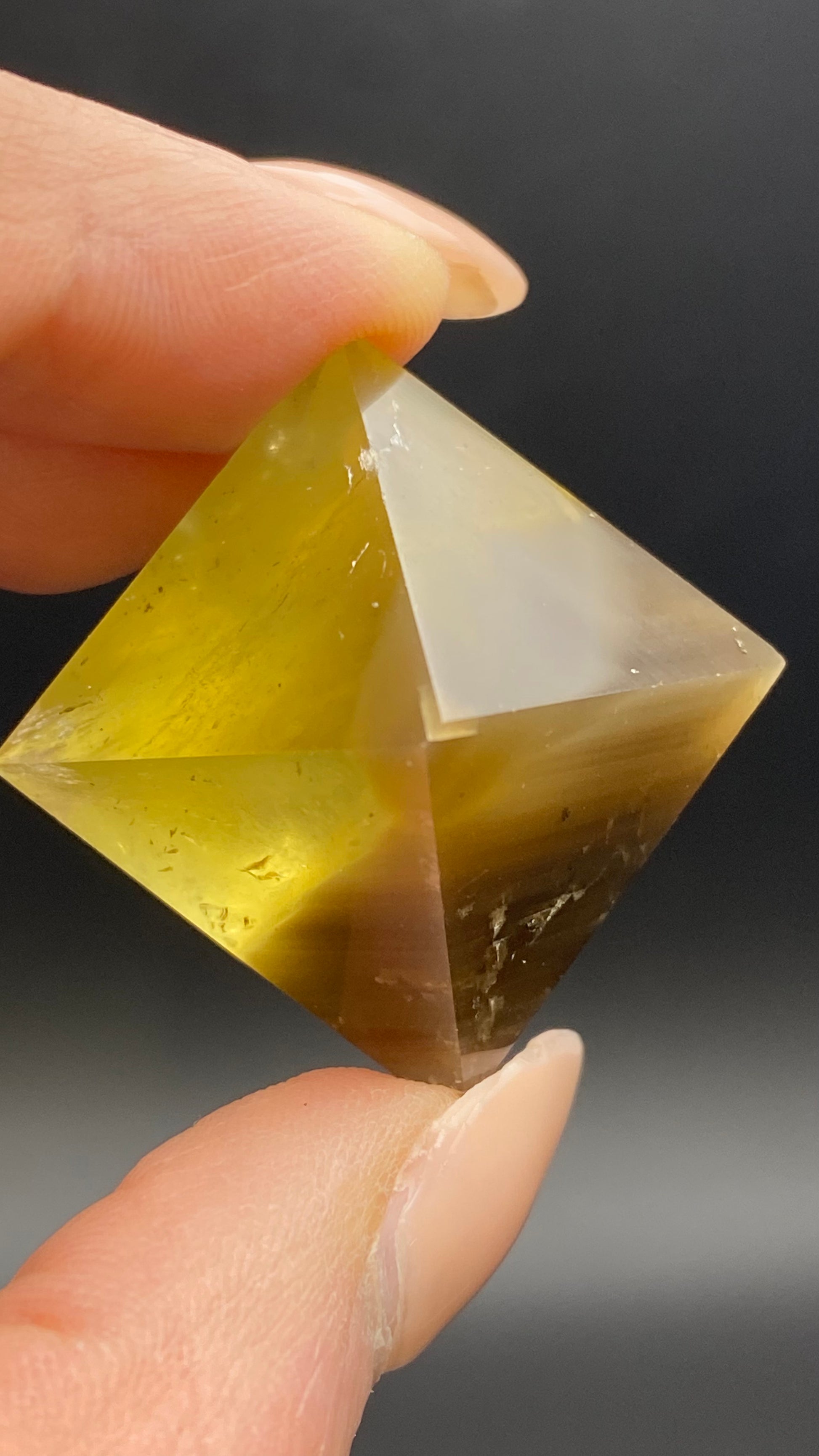 Close-up of a hand holding The Crystalary's Fluorite Octahedron from Cave-in-Rock, Hardin Co, Illinois against a dark background. The yellow-green quartz crystal, shaped into a double pyramid (octahedron) and reminiscent of hand-polished fluorite, appears slightly translucent with natural inclusions within the crystal structure.