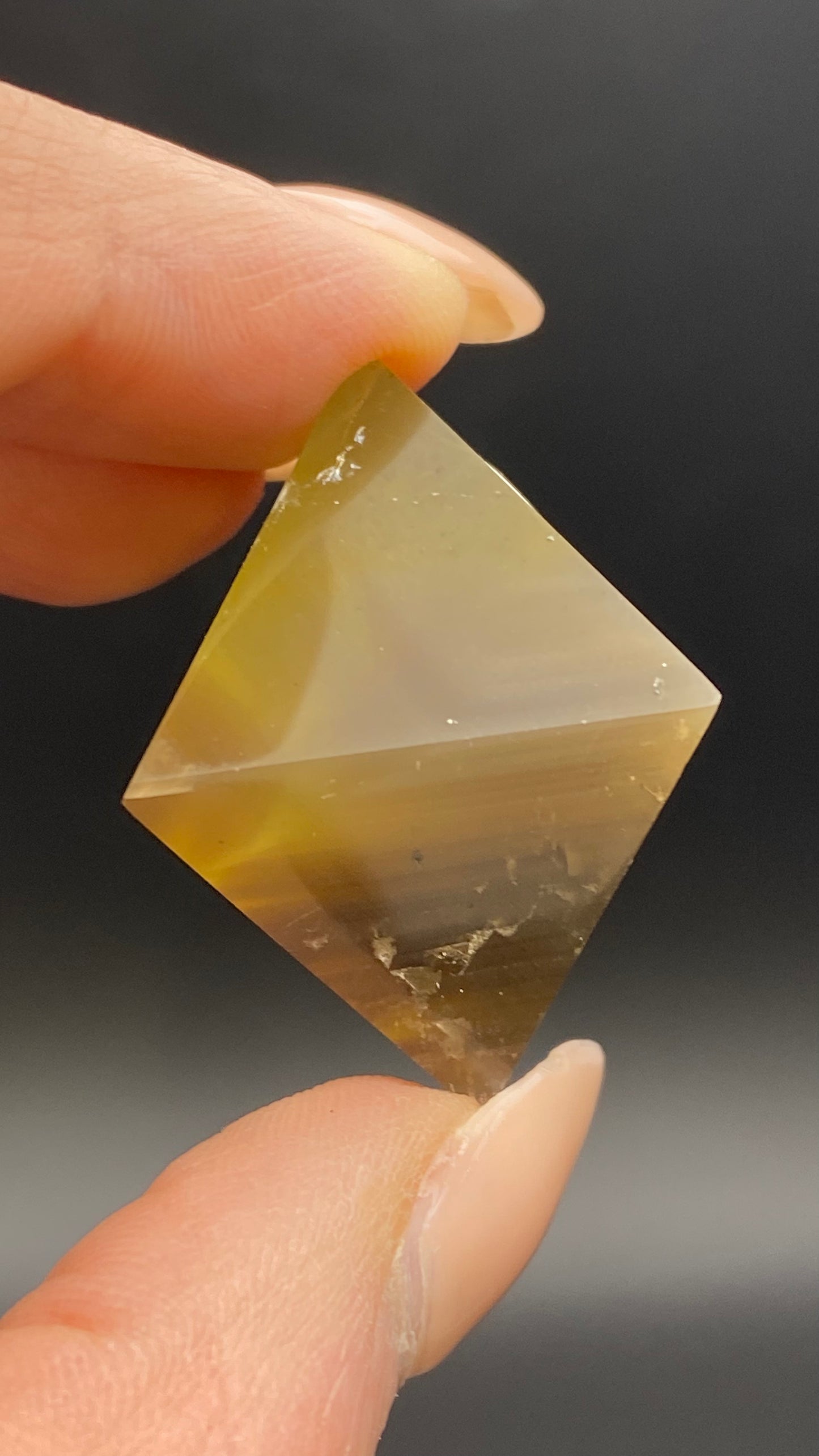 Close-up image of a hand holding a polished yellowish-brown Fluorite Octahedron from Cave-in-Rock, Hardin County, Illinois, reflecting The Crystalary's quality craftsmanship. The stone displays smooth surfaces with visible gradients of color and a reflective sheen against a dark background.