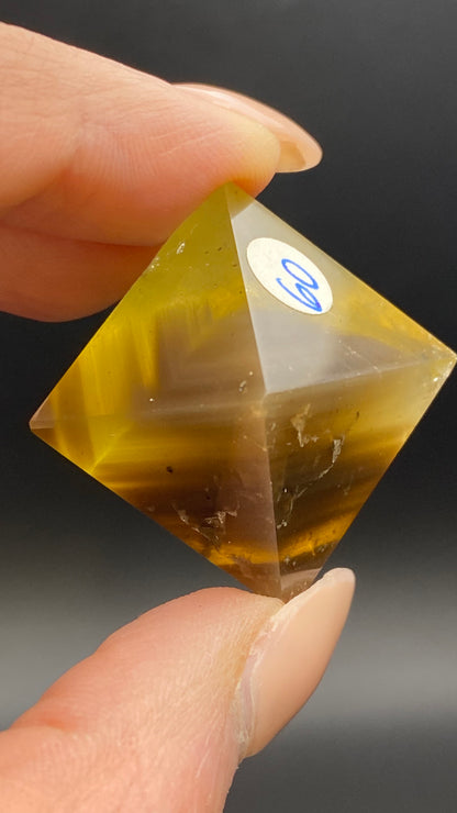 A close-up photo showcases a person's hand holding a Fluorite Octahedron from Cave-in-Rock, Hardin Co, Illinois, with yellow and brown stripes. The Crystalary's hand-polished gemstone features several layers and a small label with the number "29" on one of its faces.