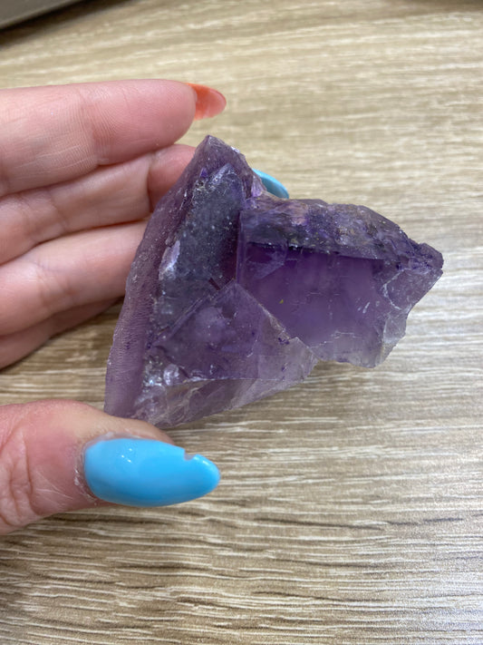 A hand with blue-painted nails is holding a rough, unpolished purple amethyst crystal from The Crystalary's LIVE- cactusoddities- 9/19/24 & 9/26/24 collection against a wooden surface background. The crystal, reminiscent of treasures from Cave in Rock, displays various shades of purple and has jagged edges.