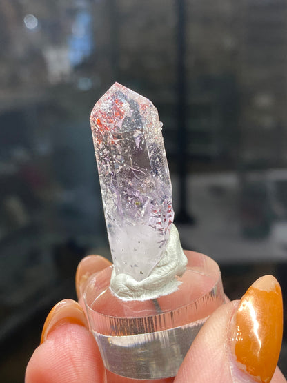 A person holds The Crystalary's "Harlequin Quartz" from the Streep Mine, Goboboseb, Namibia (sku3006)—a clear quartz crystal with red and white inclusions of lepidoicrocite and hematite. It is elegantly displayed on a transparent stand to accentuate its charm. The person's vibrant orange nails add a splash of color to the softly blurred background.