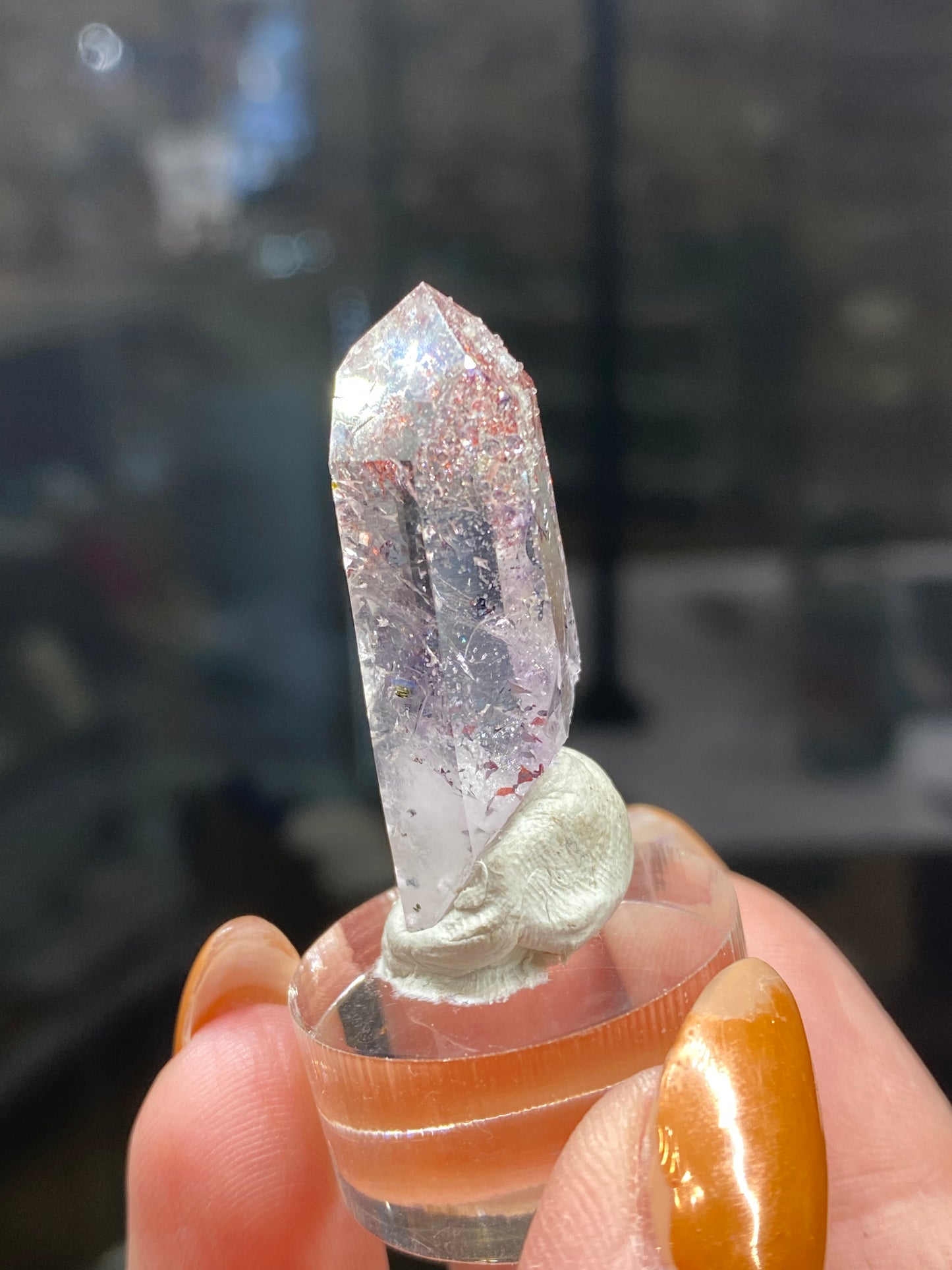 A close-up of a Quartz, Lepidocrocite, Hematite, and Analcime crystal—known as "Harlequin Quartz" from Streep Mine, Goboboseb, Namibia—is presented upright on a clear stand. The background is blurred while a hand with brownish-orange painted nails holds the stand. Product code: sku3006 by The Crystalary.