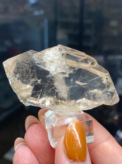 A hand with an orange manicure holds a Quartz, var. Faden from The Crystalary, positioned on a clear stand. Its large, translucent angular form and rough, reflective facets create a stunning contrast against the blurred dark background, evoking the treasures of Mont Blanc Massif's northwest flank in Auvergne-Rhône-Alpes, France (sku3008).