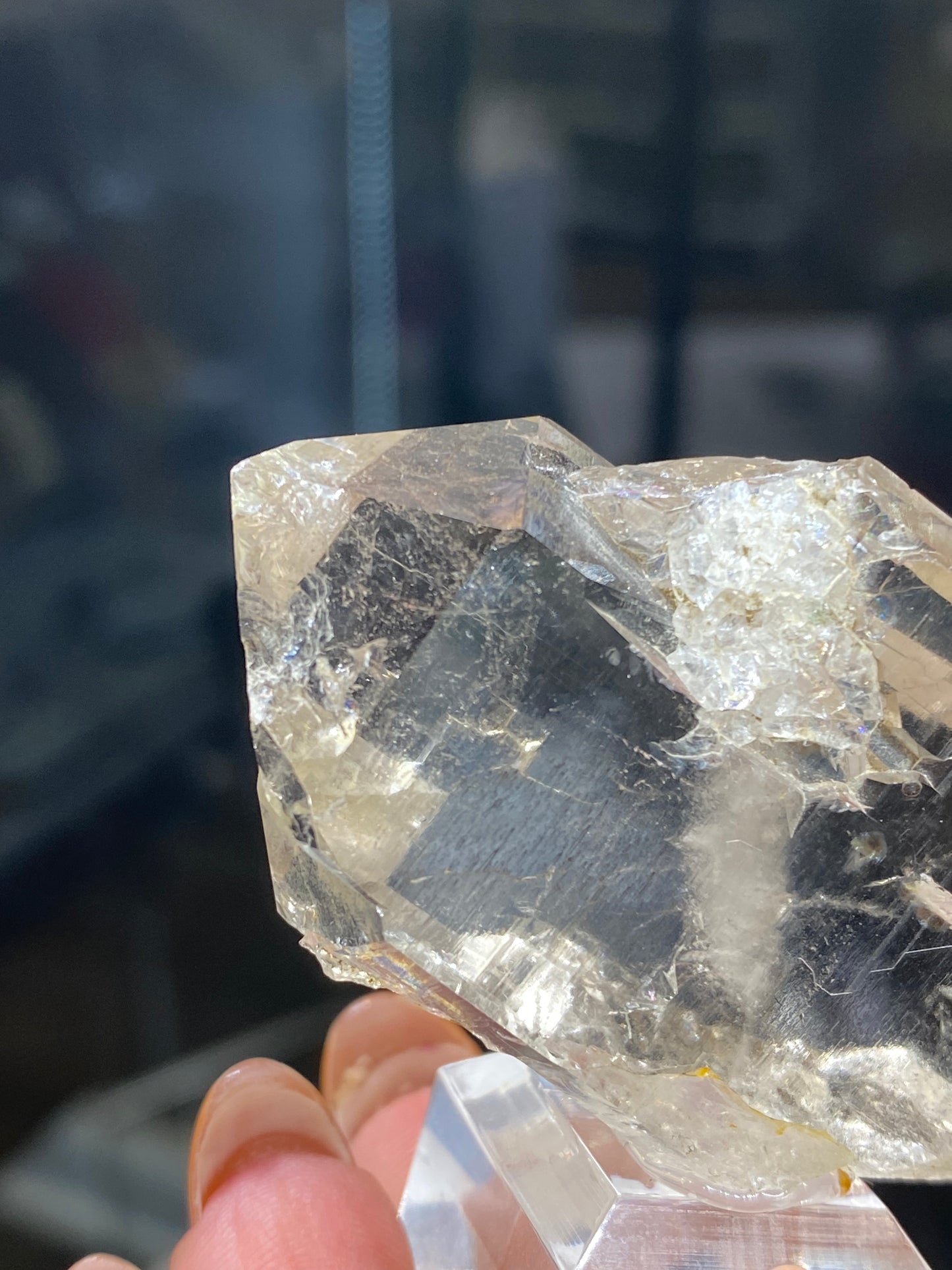 A close-up of a person's hand holding a translucent, angular quartz crystal from The Crystalary, known as Quartz, var. Faden (sku3008), with a prism-like shape. The background is blurred, emphasizing the shiny surface and intricate internal patterns of the mineral that are reminiscent of formations found on the northwest flank of Mont Blanc Massif in Auvergne-Rhône-Alpes, France.