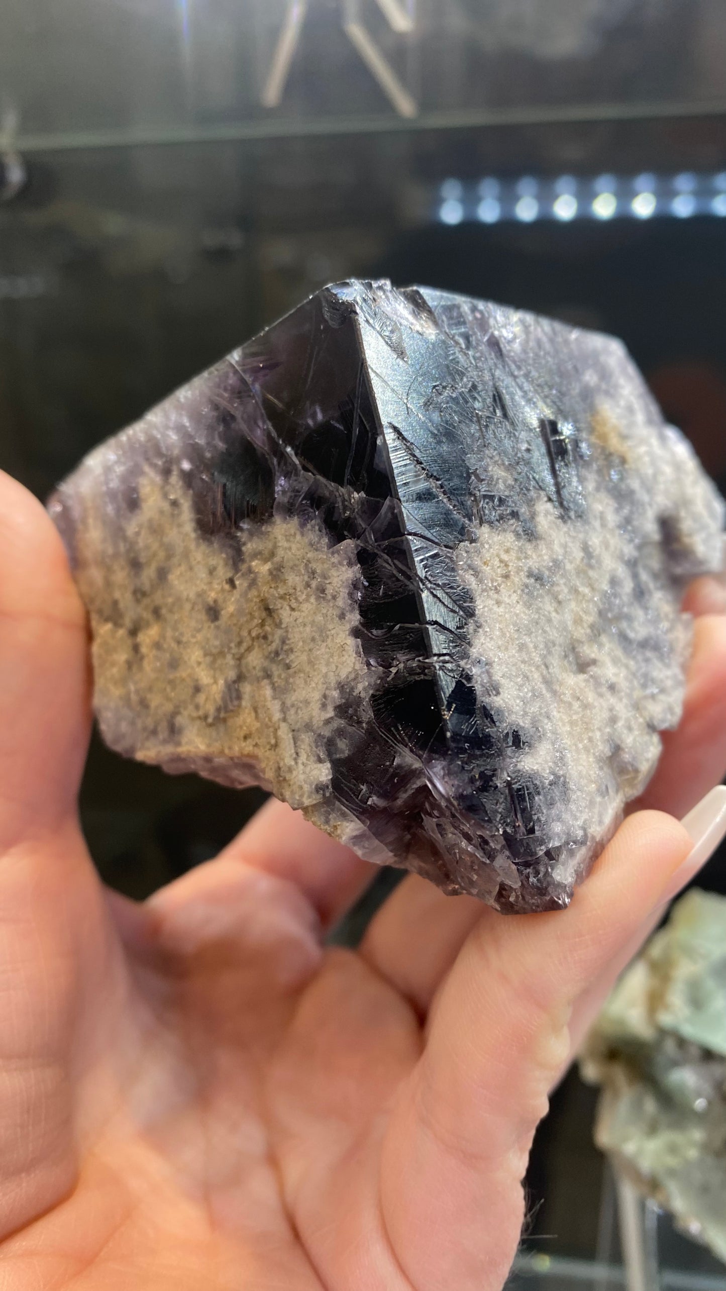 A hand holds a Fluorite - Purple Rain Pocket crystal from The Crystalary, featuring a rough, partially translucent surface. Sourced from the Lady Annabella Mine in Eastgate, Weardale, Co. Durham, England (sku 3020), this jagged-textured stone exhibits light chalky areas and displays a deep purple hue against a softly blurred background.