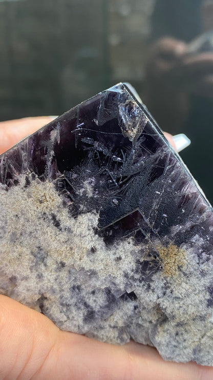 Close-up of a hand holding the Fluorite- Purple Rain Pocket crystal from The Crystalary, showcasing a blend of dark purple and light gray hues. The surface features a rugged texture with distinct natural striations and formations, similar to those found in the renowned Lady Annabella Mine in Eastgate, Weardale, Co. Durham, England. (Product SKU: 3020)