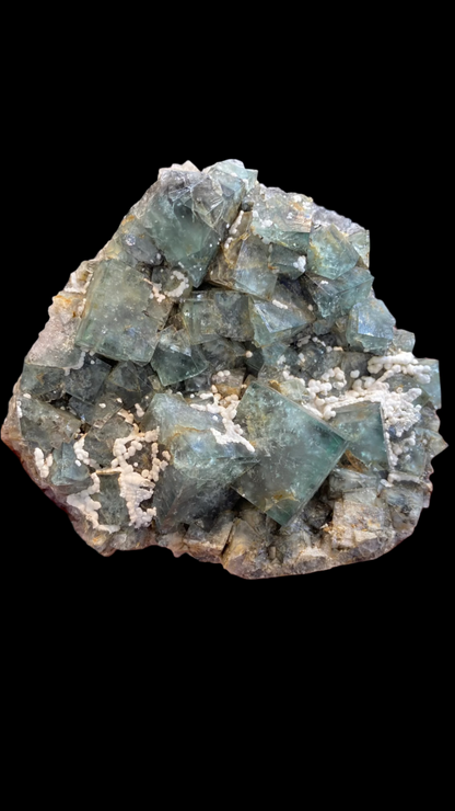 A hand holding a cluster of cubic green fluorite crystals, partially adorned with small white calcite deposits. In the background, a reflective surface and a blurred figure mirror the allure of The Crystalary's Fluorite from the enchanting Fairy Holes Pocket at the Lady Annabella Mine (sku 3021).