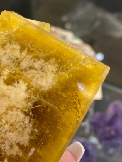 A close-up of the Fluorite- Honey Bear Pocket, a translucent, golden honey-yellow crystal with white, fibrous inclusions from The Crystalary. It is delicately held between fingers against a blurred background that suggests vibrant hues reminiscent of treasures from the Diana Maria Mine in Frosterley, Weardale, Co. Durham, England.