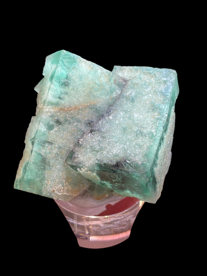 A hand displays a rough, square-shaped Fluorite - Minty Cheesecake Pocket crystal from The Crystalary, featuring visible cracks and a shimmering surface. This gem, sourced from the Lady Annabella Mine in Eastgate, Weardale, Co. Durham, England (sku 3022), stands out against the dark backdrop like a hidden treasure.