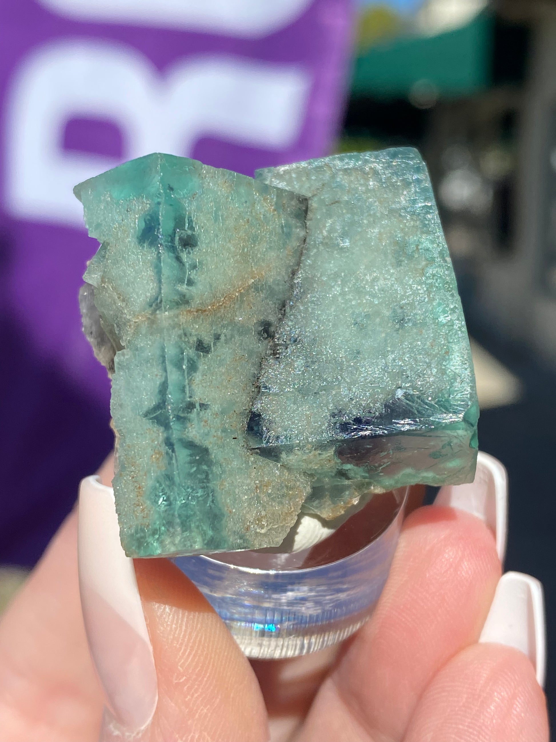 A close-up image showcases a hand holding The Crystalary's "Fluorite- Minty Cheesecake Pocket" from the Lady Annabella Mine in Eastgate, Weardale, Co. Durham, England (SKU 3022), against a softly blurred outdoor background. The crystal's visible layers and slightly translucent appearance beautifully reflect the sunlight.