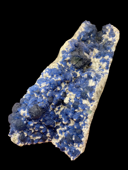 A hand holds a mineral specimen from The Crystalary, featuring vibrant blue crystals on a pale rock matrix, reminiscent of Fluorite from No. 4 Mine, Huanggang, Chifeng City, Inner Mongolia, China. The slightly blurred background showcases various other minerals and textured surfaces from the Huanggang region.