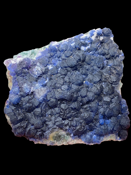 A hand holds a vibrant specimen of Fluorite from The Crystalary, sourced from the No. 4 Mine in Huanggang, Chifeng City, Inner Mongolia, China (SKU 3037). The mineral features a textured surface with deep blue and purple crystalline formations that resemble Prussian blue. It sparkles under light, showcasing its intricate patterns.