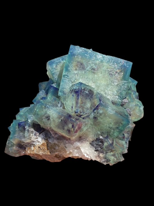 A hand holds a cluster of vibrant blue and green Fluorite from The Crystalary's Northern Lights Pocket collection, sourced from Diana Maria Mine in Frosterley, Weardale, Co. Durham, England (SKU 2028), featuring sharp geometric shapes. The background reveals a sunny outdoor street scene with blurred buildings.