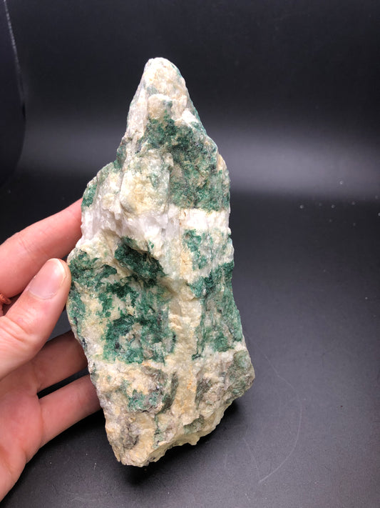 A hand holds "AUCTION Mariposite- Carson Hill, Calaveras Co, CA" by The Crystalary against a dark background. The rugged surface of the large, jagged rock reveals patches of green Mariposite intertwined with striking white quartz banding, creating an eye-catching contrast.