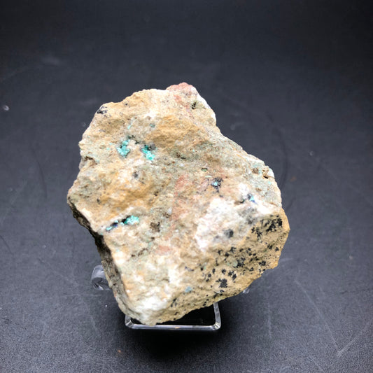 A rough, light-colored mineral rock with small specks of green and black sits on a dark surface. This Theisite specimen, sourced from Tucker's Tunnel in Tuckerville, Hinsdale Co., Colorado by The Crystalary, has a slightly uneven texture and irregular shape, positioned on a small transparent stand for display.