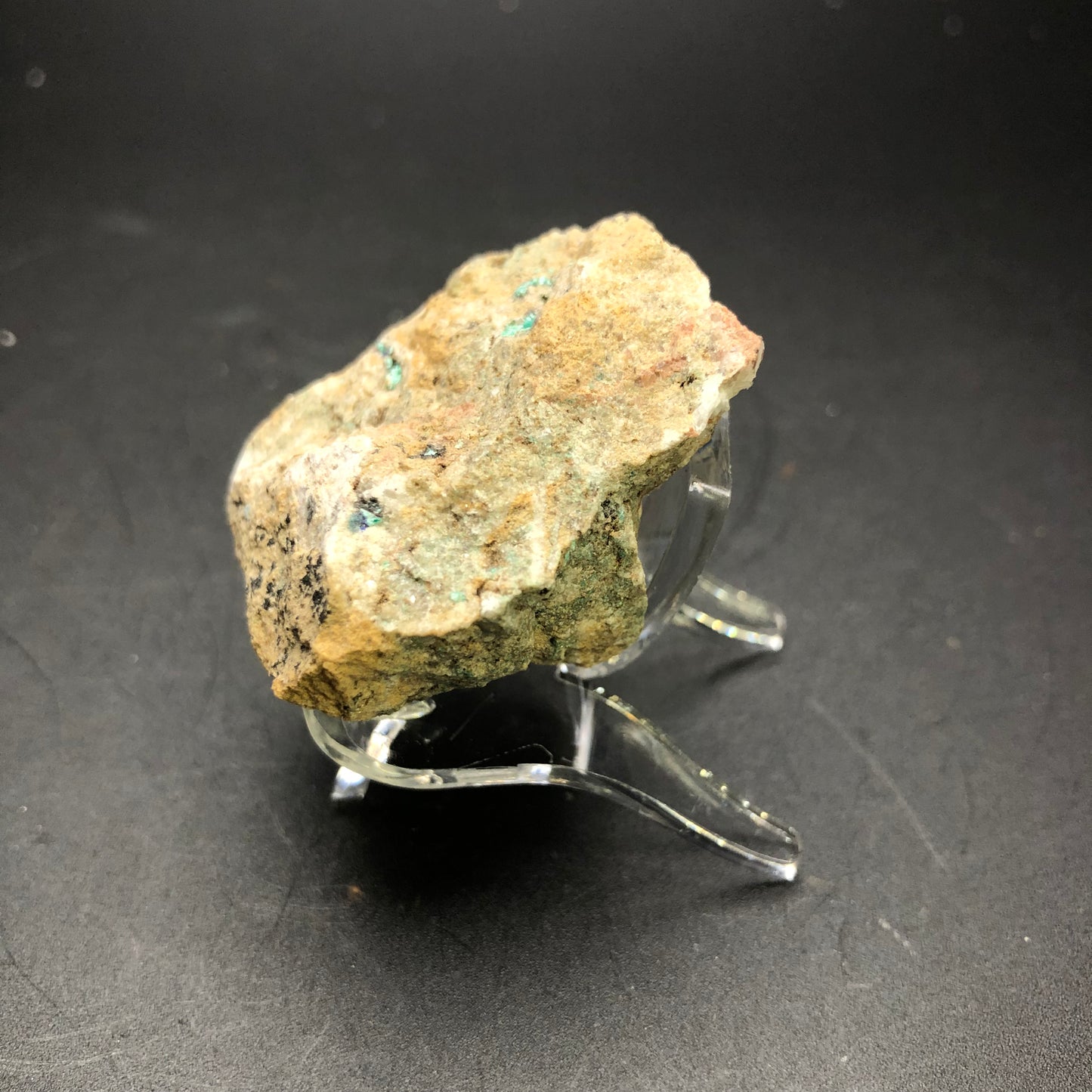 A rough, earthy-colored mineral rock with speckles of greenish and bluish hues, identified as AUCTION Theisite from Tucker's Tunnel, Tuckerville, Hinsdale Co., Colorado, USA by The Crystalary, is displayed on a clear acrylic stand against a black background. The rock has an uneven surface texture with visible jagged and irregular edges.