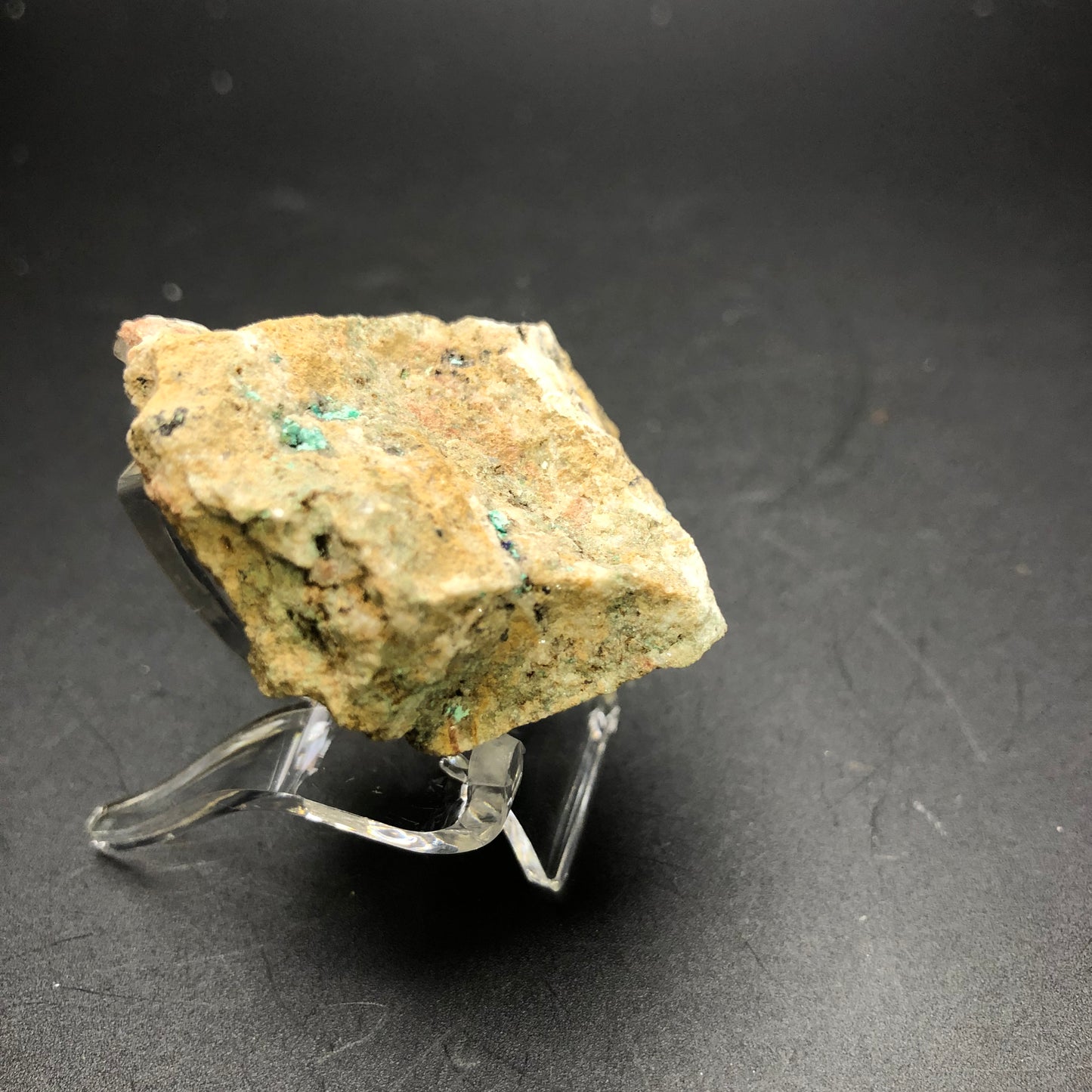 A small and coarse-textured specimen of Theisite from The Crystalary, featuring green and tan patches, is displayed on a transparent stand against a black background; sourced directly from Tucker's Tunnel in Tuckerville, Hinsdale Co., Colorado.