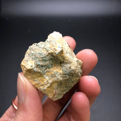 A hand holding a roughly textured rock with shades of beige and green, identified as an AUCTION Theisite specimen from The Crystalary's Tucker's Tunnel in Tuckerville, Hinsdale Co., Colorado, USA. The background is dark and out of focus, making the rock the central point of interest.