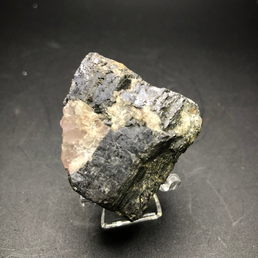 A close-up of "AUCTION Galena- Cave-in-Rock, Hardin County, Illinois, USA" from The Crystalary against a dark background. The mineral sample displays a mix of dark gray and pinkish hues with a metallic sheen and jagged texture. It is showcased on a small, clear stand along with striking veins reminiscent of purple fluorite.