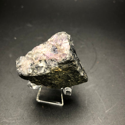 A rough mineral specimen from Cave-in-Rock sits on a small, clear stand against a black background. The predominantly dark surface features patches of translucent pinkish hues and purple fluorite, with textures that showcase its crystalline structure. This exquisite piece is labeled AUCTION Galena from Hardin County, Illinois, USA by The Crystalary.