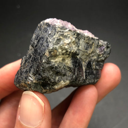 A person holds a rough, unpolished specimen of AUCTION Galena from Cave-in-Rock, Hardin County, Illinois, USA, by The Crystalary. The mineral showcases dark, textured surfaces with light purple fluorite accents on the top edge against a dark background. Its layered and jagged structure bears a striking resemblance to those found in the Cave-in-Rock mines.