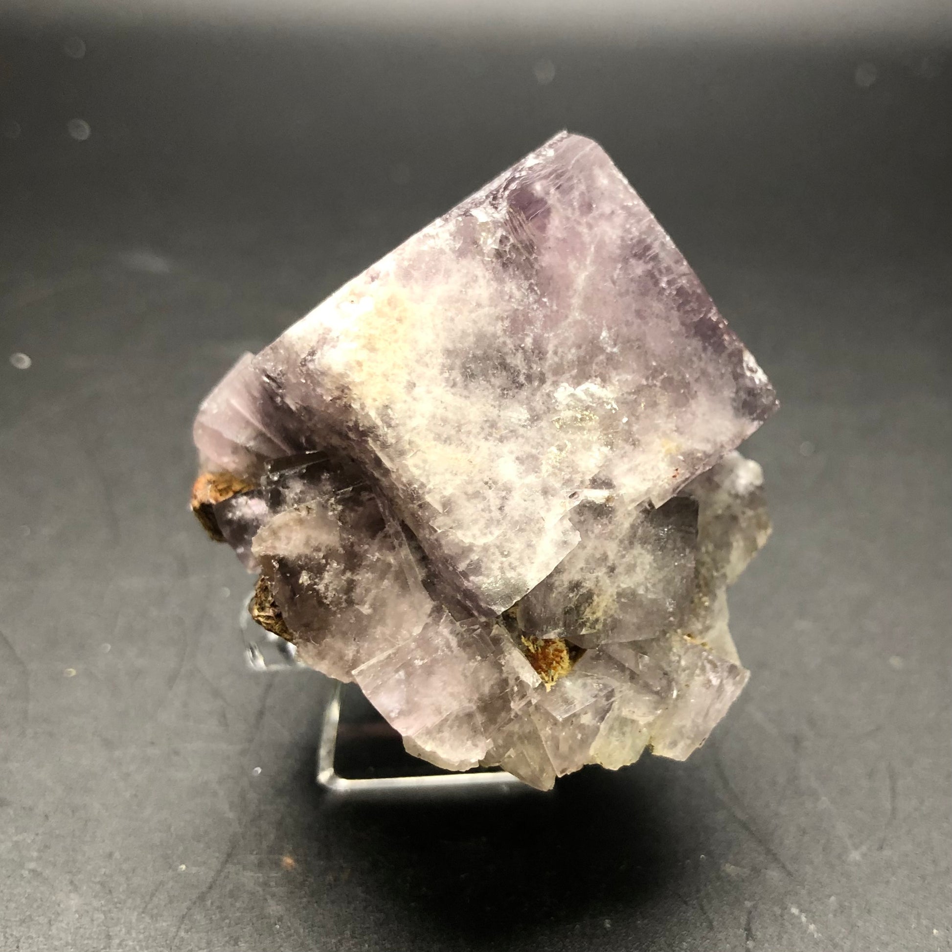 A close-up of a large cubic Purple Fluorite crystal with a metallic luster, showcasing shades of violet and light purple. This exquisite crystal from The Crystalary's AUCTION Fluorite collection, sourced from the Cromwell Pocket in Greenlaws Mine, Daddry Shield, Stanhope, County Durham, UK, is set against a dark background and stands gracefully on a small, transparent display stand.