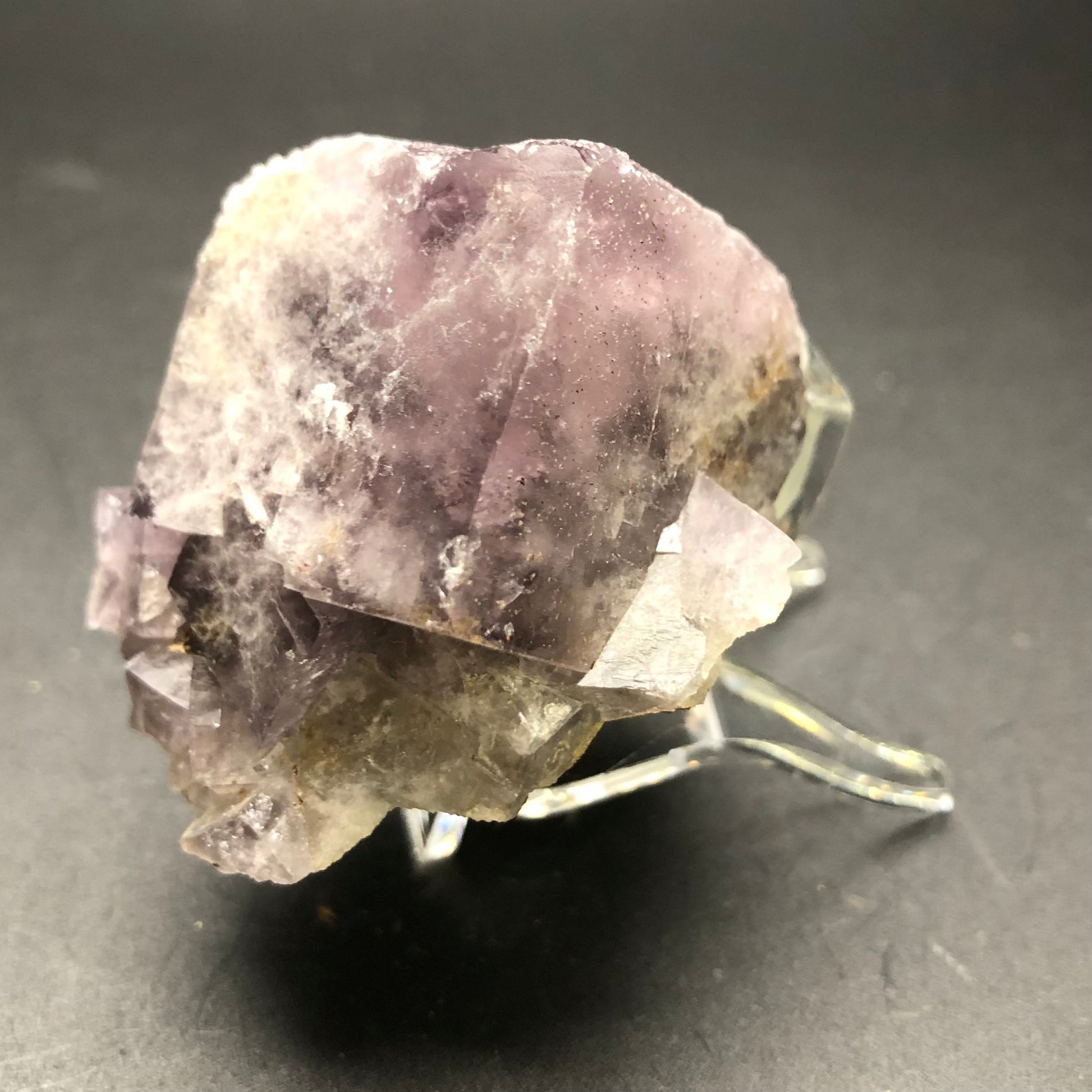 A large, rough fluorite crystal from The Crystalary's AUCTION series rests on a clear display stand against a dark background. Exhibiting shades of purple with some cloudy white inclusions, its natural, unrefined beauty and irregular shape are reminiscent of the fine specimens found in Cromwell Pocket, Greenlaws Mine, County Durham, UK.