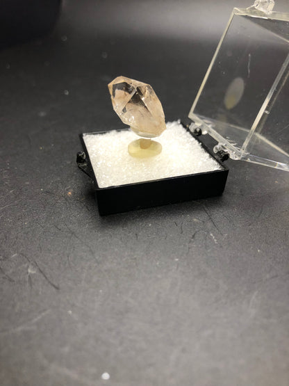 A clear, miniature crystal from The Crystalary's AUCTION Smoky Quartz collection, originating from Minas Gerais, Brazil, is displayed on a small pedestal inside a hinged black box with an open transparent lid. The black box rests on a textured black surface. This light-colored mineral is elegantly mounted on a white cushion.