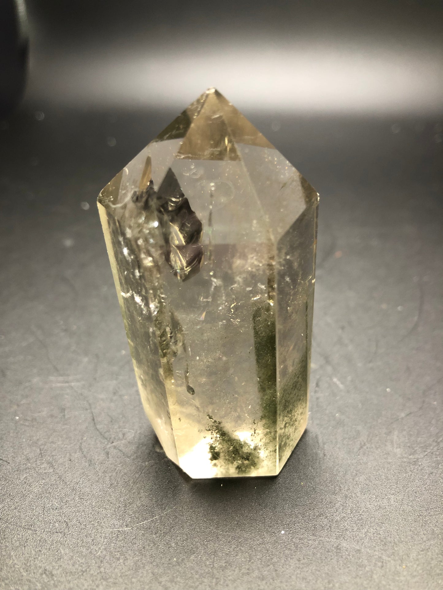 A close-up of The Crystalary's AUCTION Garden Quartz from an unknown locality, showcasing a polished, clear point with visible natural inclusions and greenish mineral deposits. Set against a dark background, the quartz features a pyramidal top, a hexagonal base, and occasional rainbow flashes that enhance its mesmerizing allure.