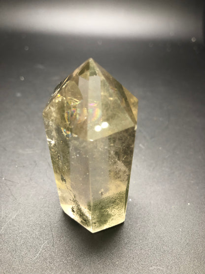 The AUCTION Garden Quartz from The Crystalary stands upright on a dark surface, with its hexagonal prism shape culminating in a pointed, pyramid-like tip. Parts of the crystal display rainbow-like reflections and slight inclusions within its structure.