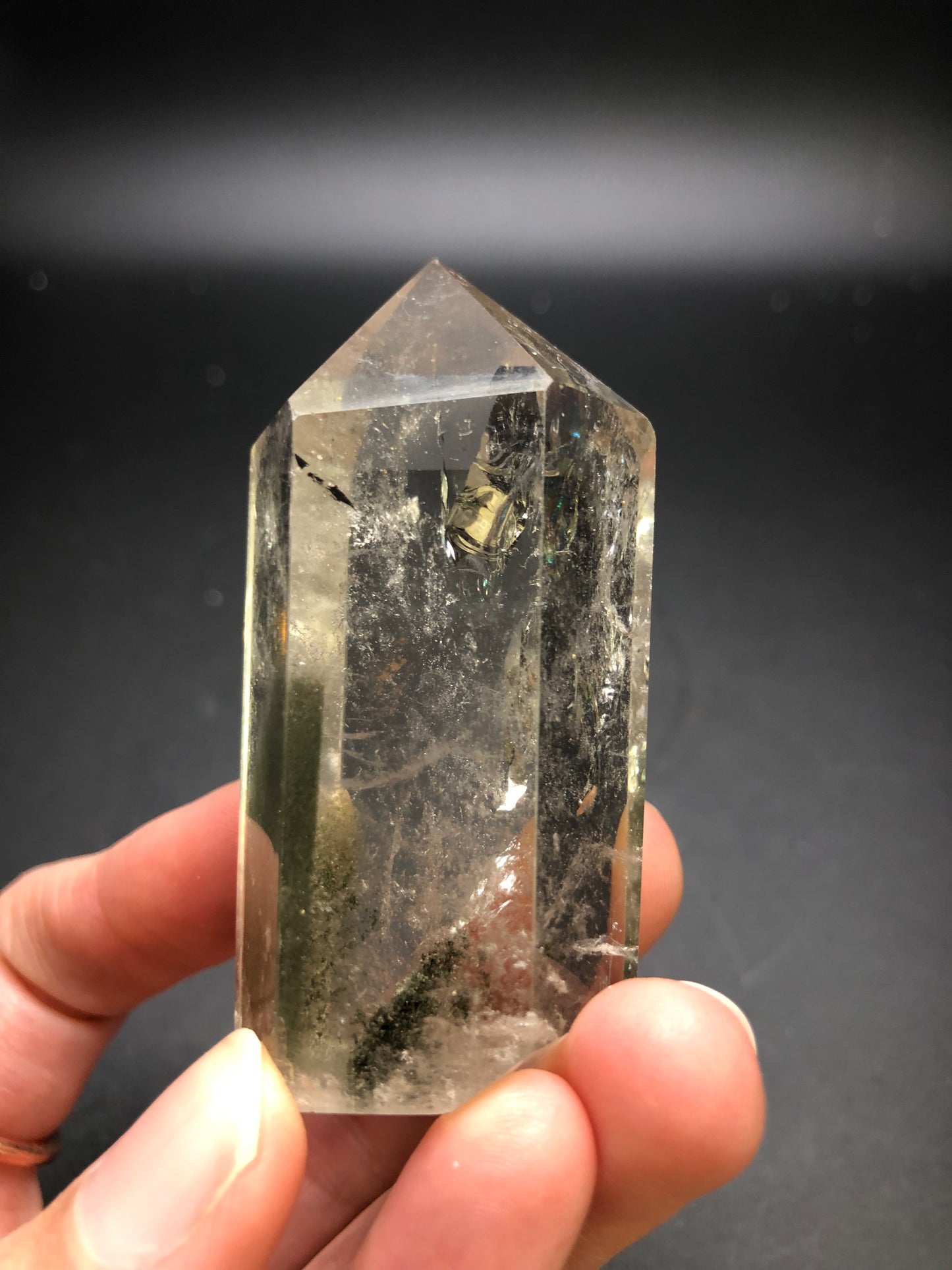 A hand is holding the AUCTION Garden Quartz from The Crystalary against a dark background, showcasing its clear, hexagonal crystal point with a pointed tip. Inside the quartz are natural inclusions, including a small metallic cube and a mesmerizing rainbow flash.