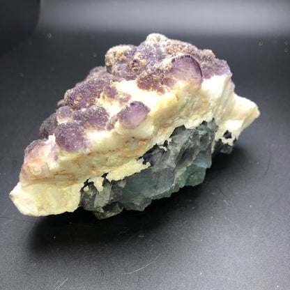 A cluster of assorted minerals from the Navidad Mine in Durango, Mexico, part of The Crystalary's AUCTION Fluorite collection, rests on a dark surface. The top features light purple formations with a rough texture reminiscent of cheesecake fluorite, while the bottom showcases greenish and translucent crystal formations. The overall structure offers a striking contrast through its various colors and textures.