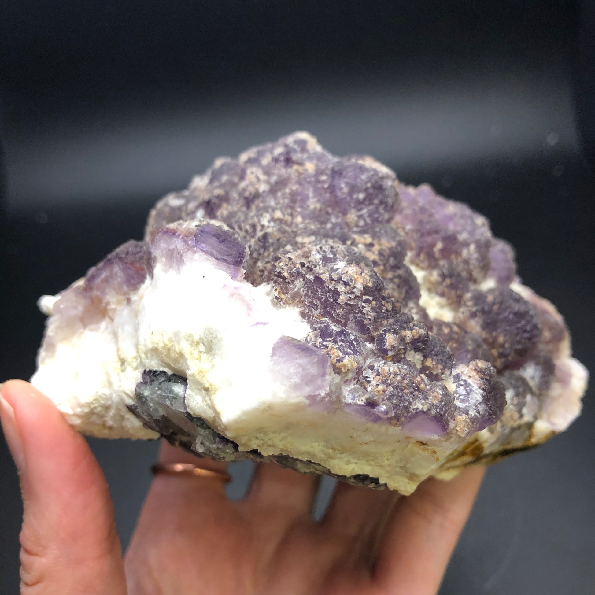 A hand holding a large, rough, purple and white mineral specimen against a dark background. This Cheesecake fluorite from The Crystalary originates from the Navidad Mine in Indé Municipality, Durango, Mexico. It features a textured crystalline surface with varying shades of purple and white, appearing somewhat glittery in patches.