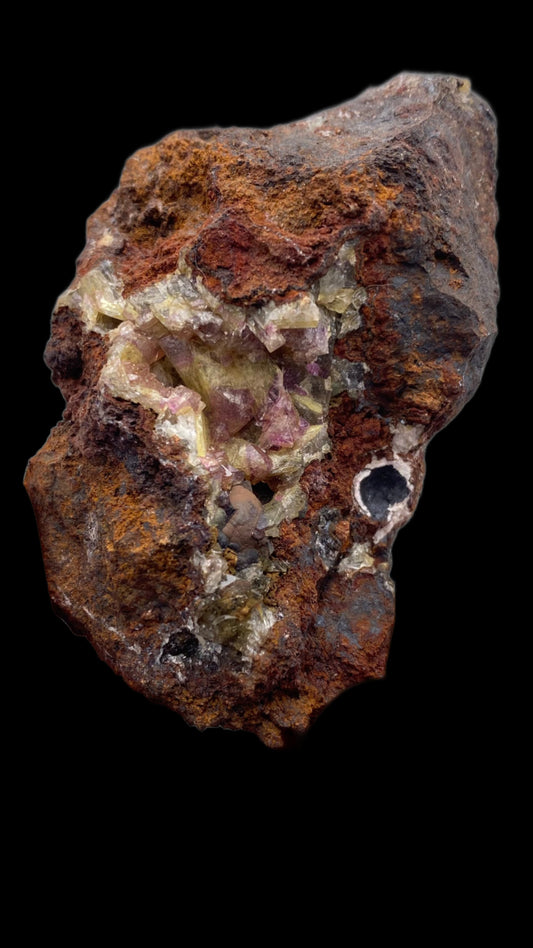 A rough, dark brown rock from The Crystalary's collection of Adamite (purple/Manganoan) from the Ojuela Mine in Mapimí, Durango, Mexico features crystalline formations with a yellowish-green hue nestled within its crevices against a black background. Some regions of the rock display small, white and slightly pinkish crystals typical of Manganoan Adamite from this renowned mine.