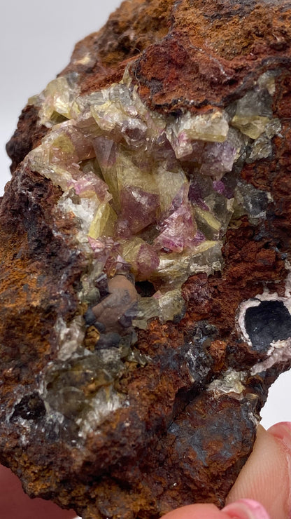 Close-up of a hand holding a rough rock specimen featuring yellow and purple crystal formations. The Adamite (purple/Manganoan) from the Ojuela Mine, Mapimí, Durango, Mexico—offered by The Crystalary—has a rugged, rusty brown exterior with a mix of other colors and crystalline structures embedded within it.