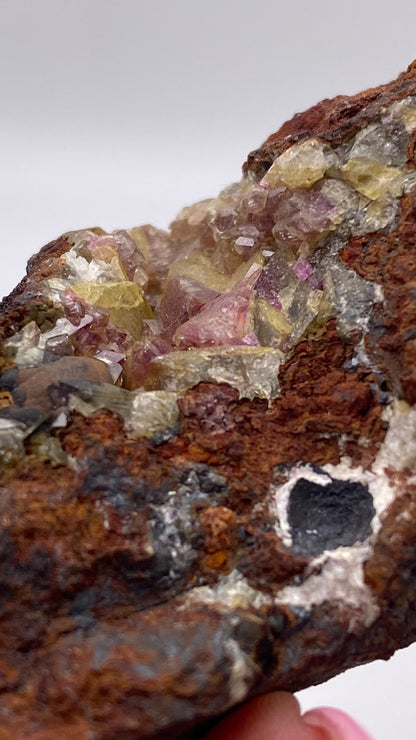 Close-up of a rough Adamite (purple/Manganoan) mineral specimen featuring clusters of small, transparent crystals in pale yellow and light purple hues on a brown, rocky matrix. The rock is irregularly shaped with a dark hole visible on one side, likely originating from the renowned Ojuela Mine in Mapimí, Durango, Mexico. Available from The Crystalary.