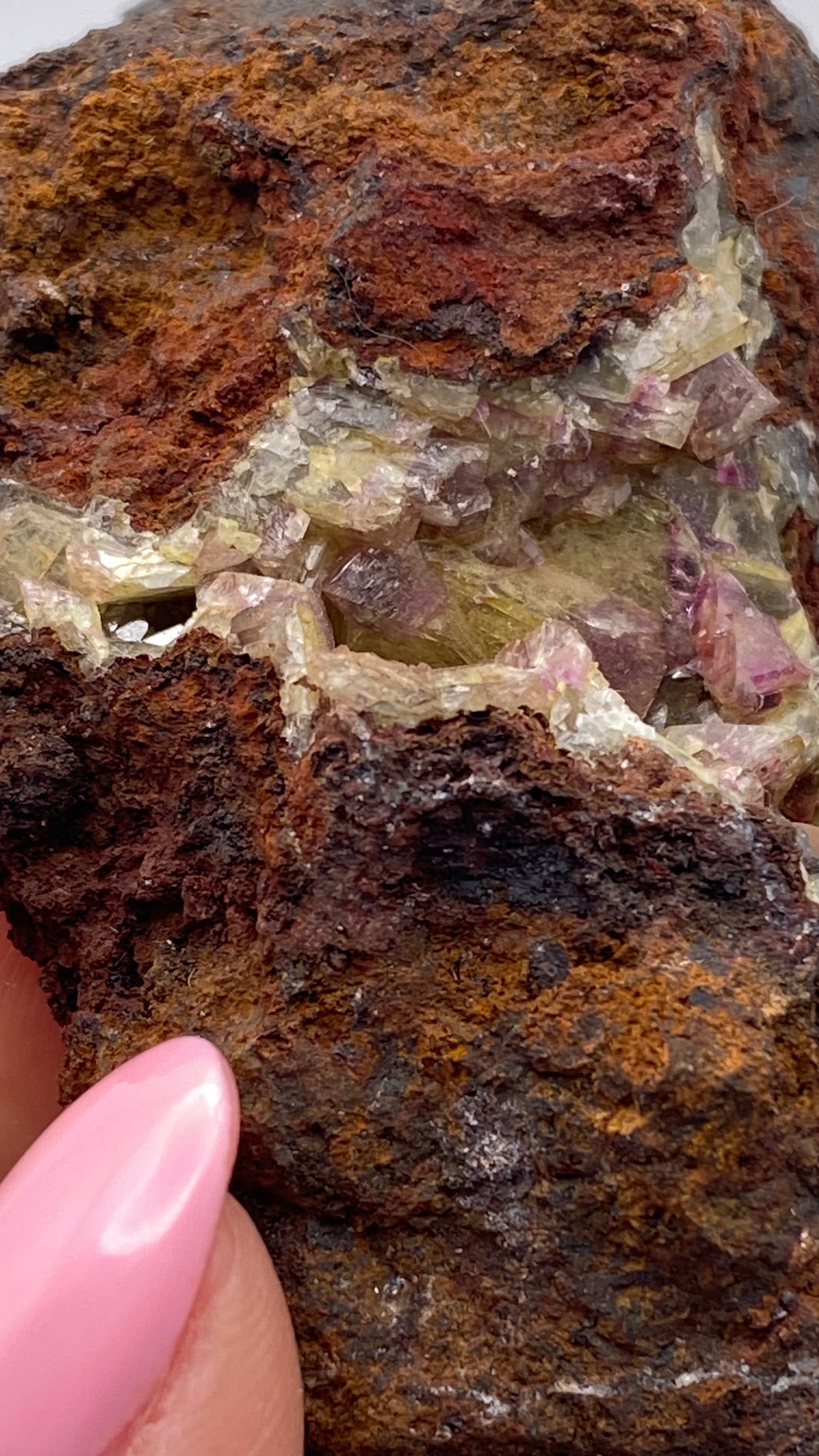 A close-up of an Adamite (purple/Manganoan) specimen from the Ojuela Mine, Mapimí, Durango, Mexico features a captivating blend of colors and textures. The layers display rust-colored and black surfaces interspersed with translucent crystalline sections that reveal green and purple mineral formations. A thumbnail with pink nail polish holds this exquisite piece from The Crystalary.