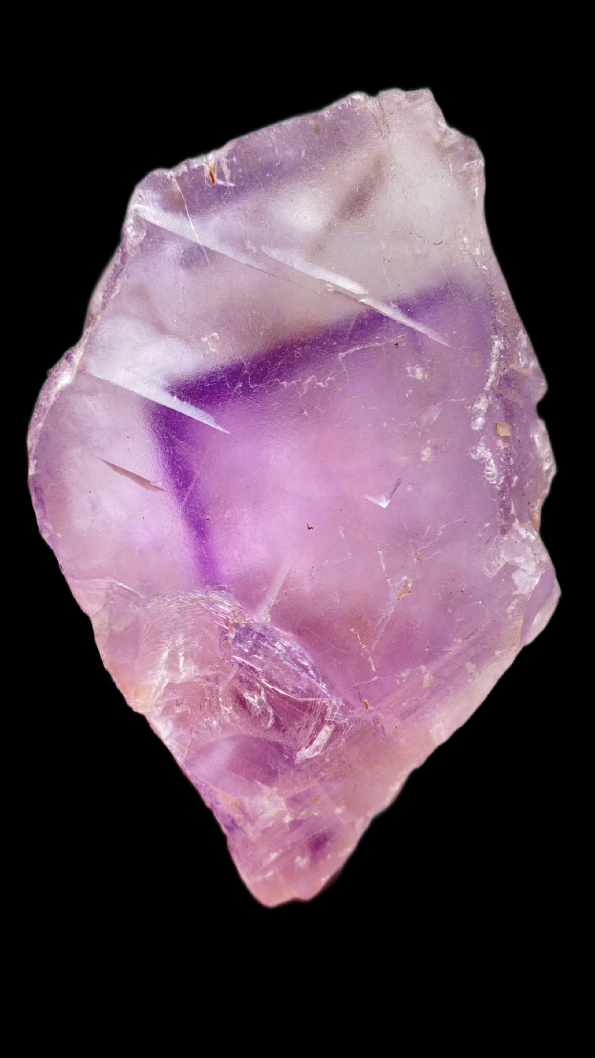 A close-up image of an unpolished Fluorite gemstone from the Cave-In-Rock region in Lead Hill, Hardin County, Illinois. This stone, offered by The Crystalary, displays a range of captivating purples from light lavender to deep violet and features visible fractures and surface irregularities. The background is solid black.